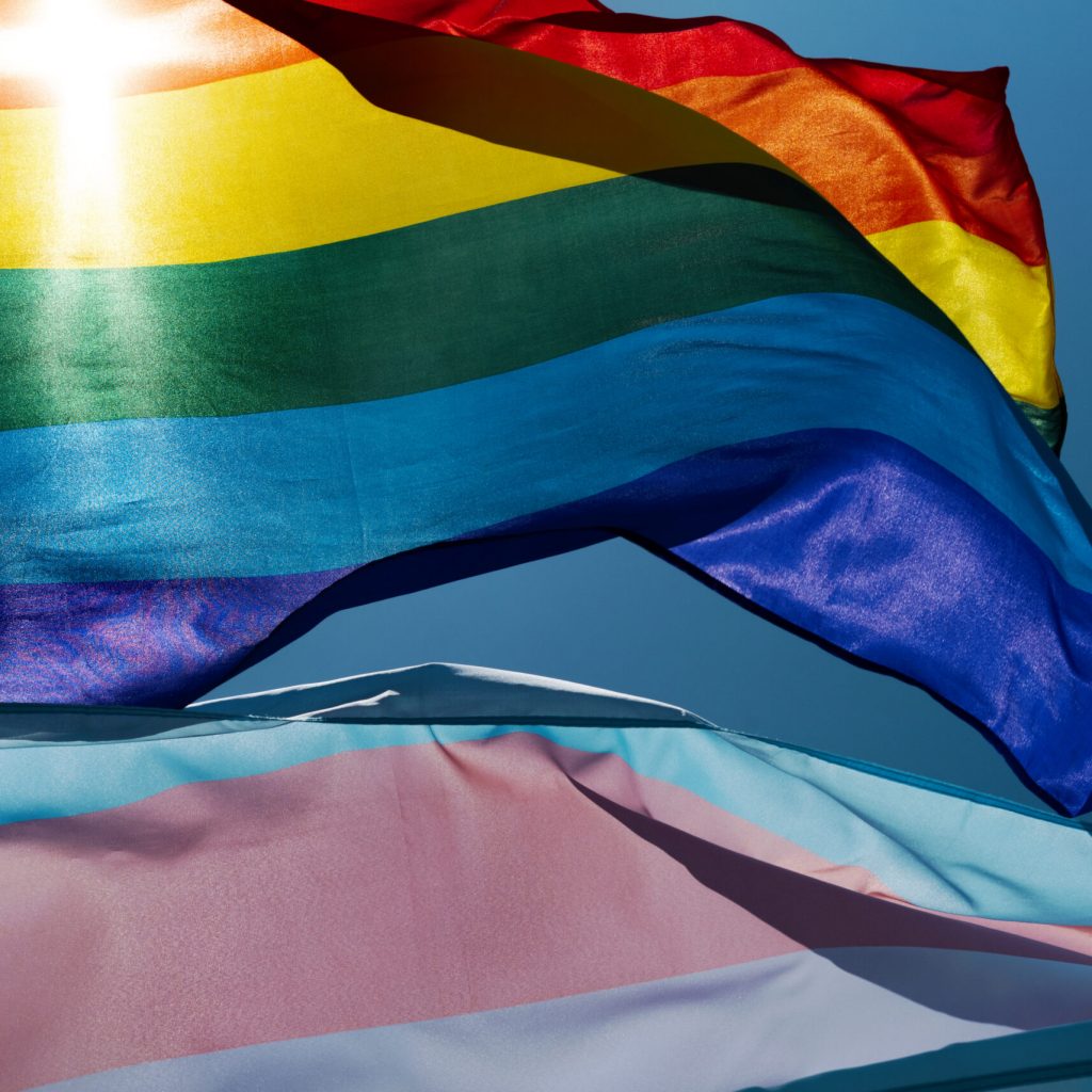 closeup of a gay pride flag and a transgender pride flag waving on the blue sky, moved by the wind, with the sun in the background